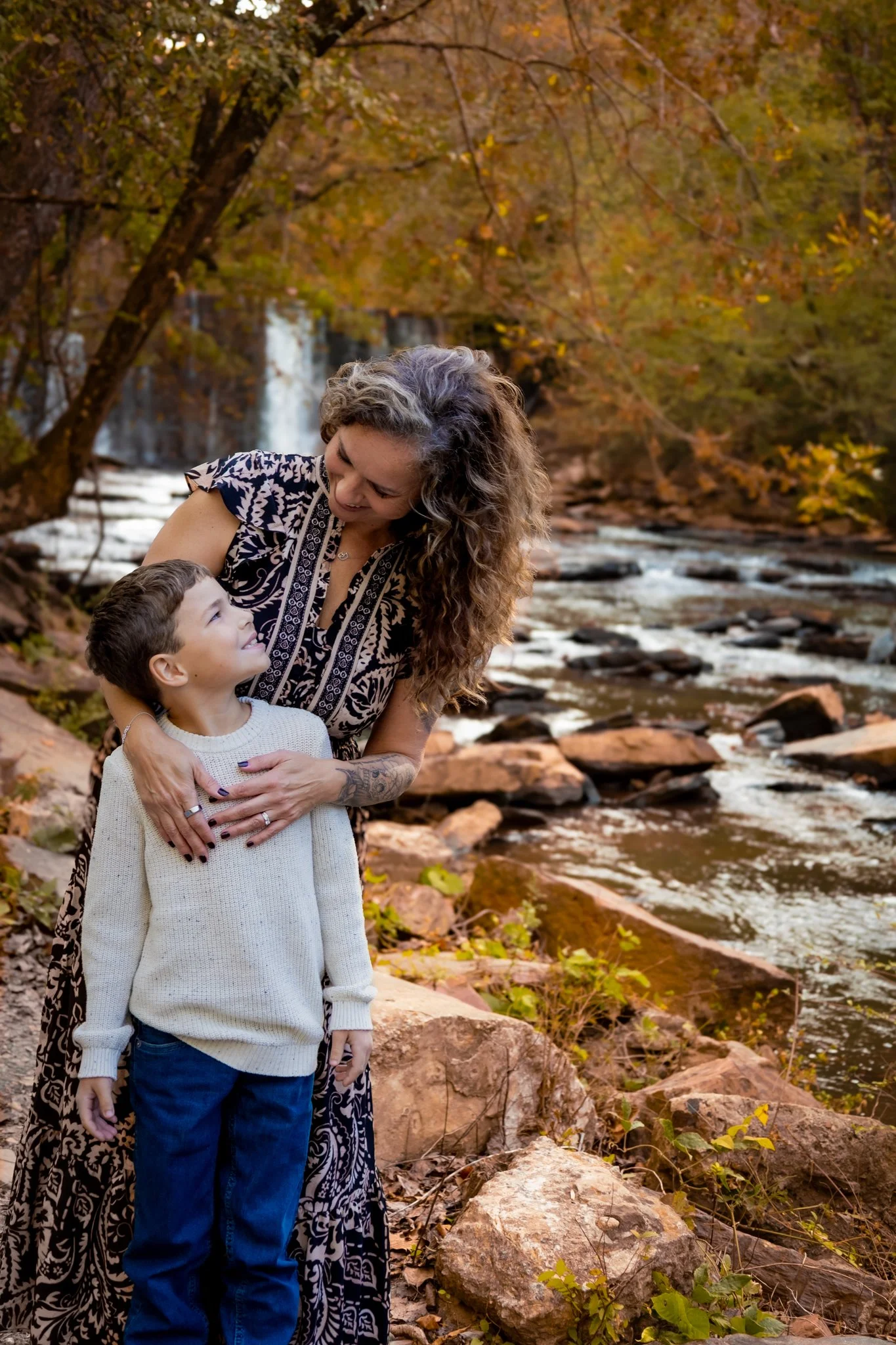 A woman with long curly hair and tattoos on her arm is smiling and embracing a young boy by a river in a forest during autumn. The background features trees with orange and yellow leaves and a waterfall.