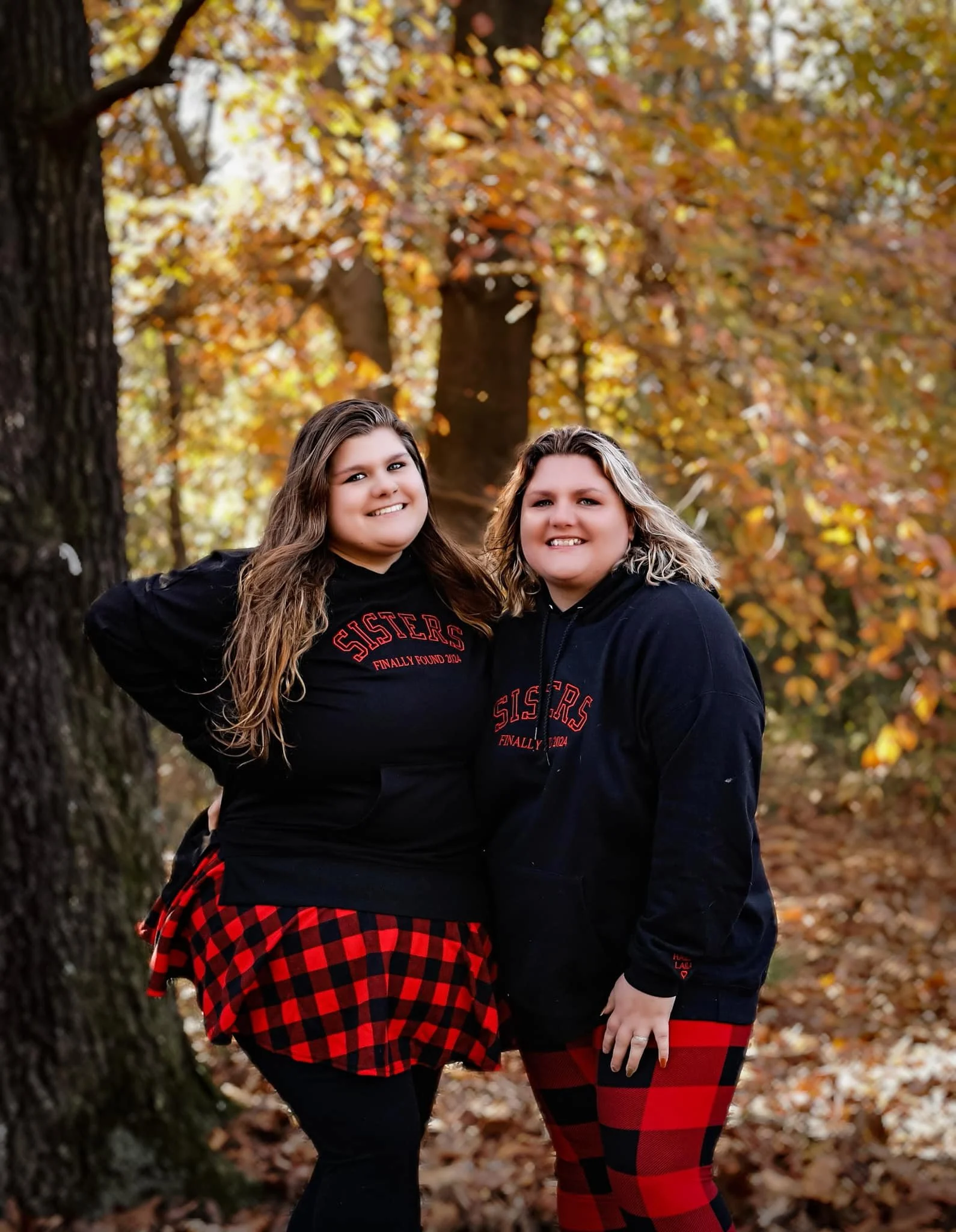 Two women standing outdoors on a fall day with orange and yellow leaves in the background, smiling and wearing matching black hoodies with red text and red plaid skirts or leggings.