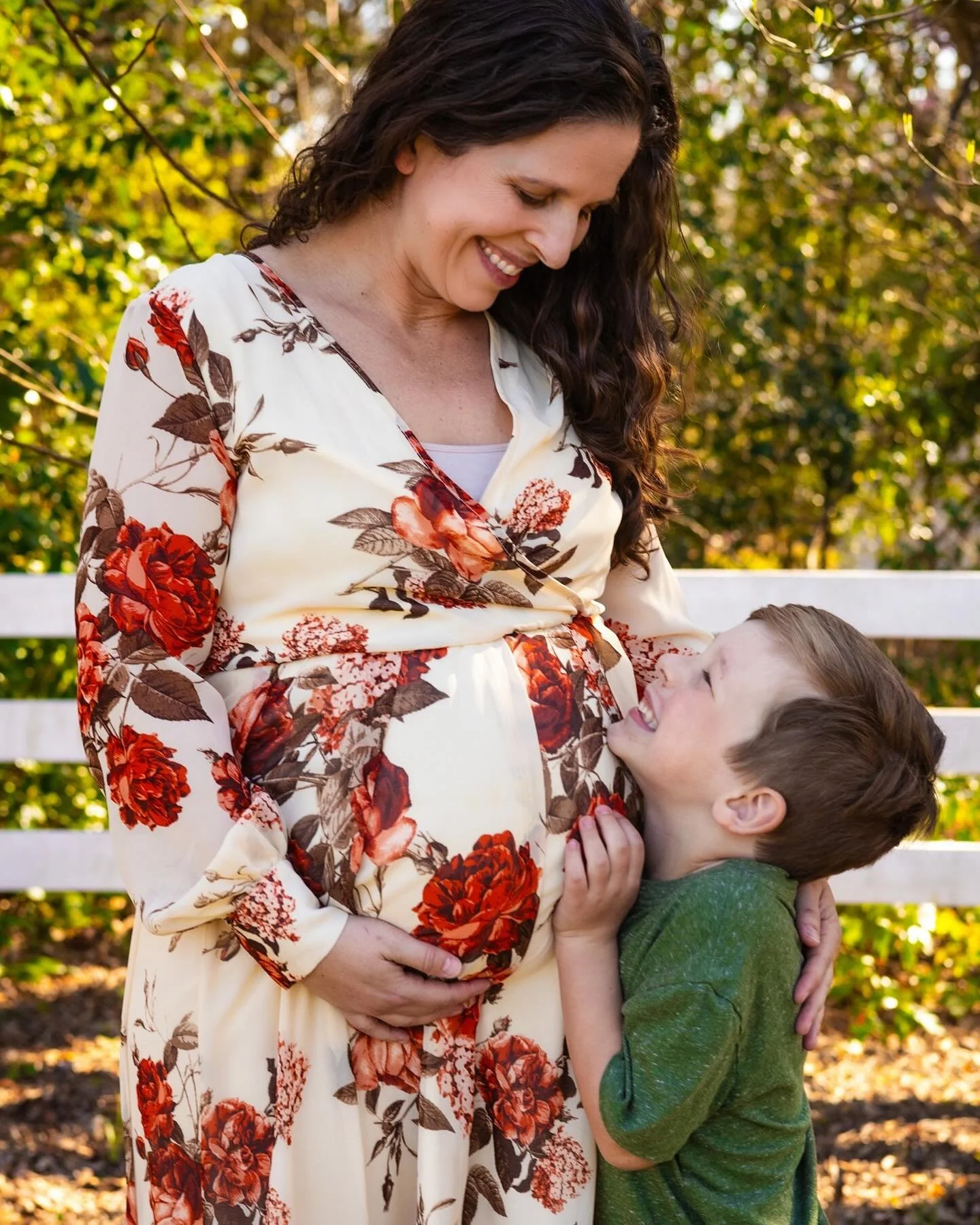 A pregnant woman in a floral dress smiling at a young boy hugging her belly outdoors.