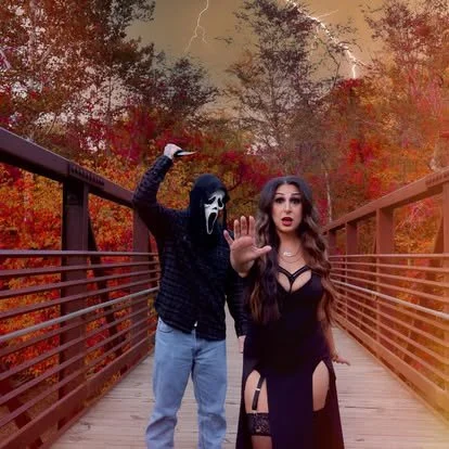 A woman in a black dress with cutouts stands on a bridge, holding up her hand as if to stop someone. Next to her, a person in a mask resembling the Scream movie character and a hoodie stands with one arm stretched out on the bridge railing. The background features autumn-colored trees.