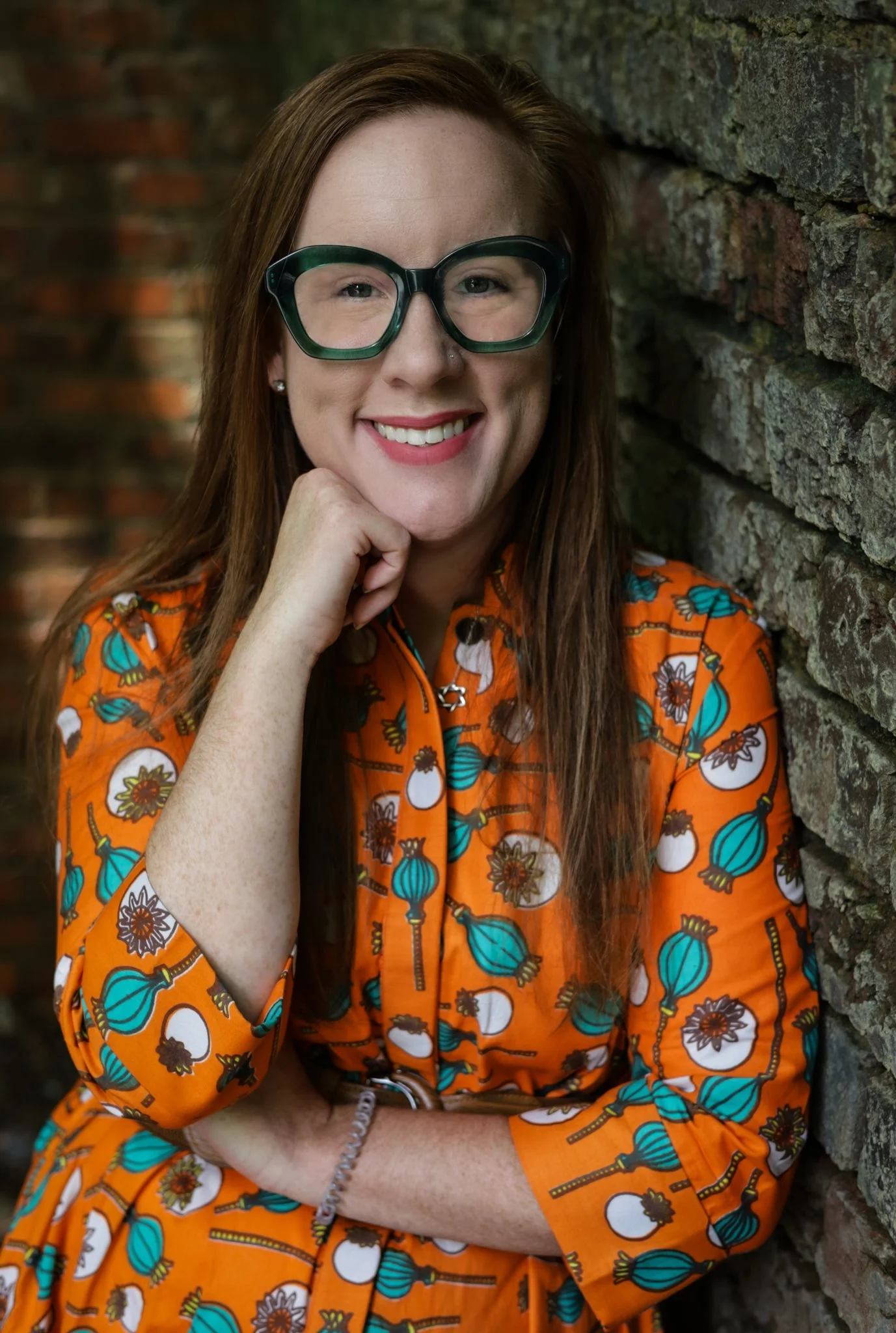 A woman with red hair, glasses, and freckles smiling, wearing an orange dress with a floral and vegetable pattern, leaning against a brick wall.