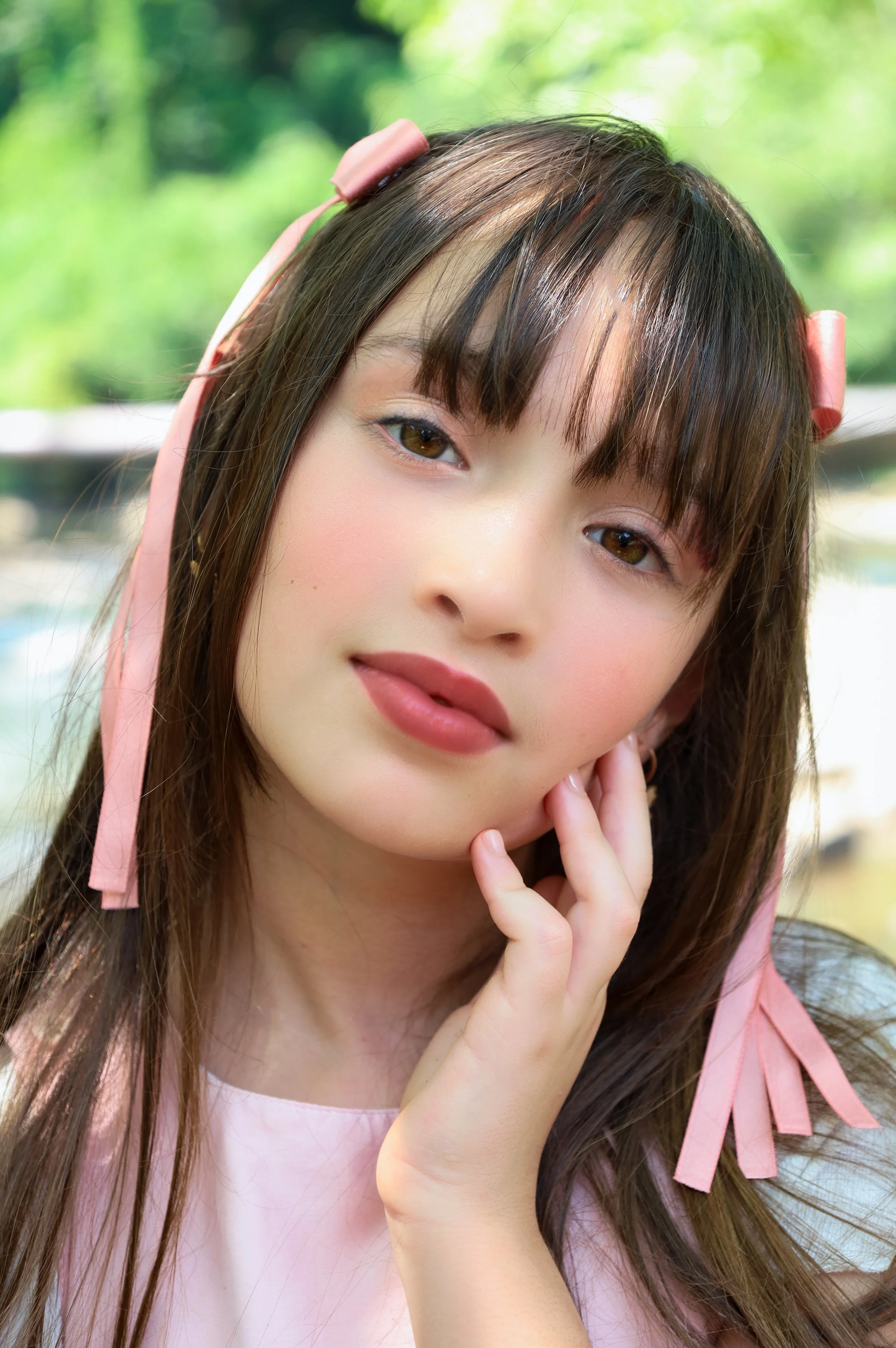 A young woman with long brown hair, pink hair ribbons, and wearing a light pink top, outdoors with green trees in the background.