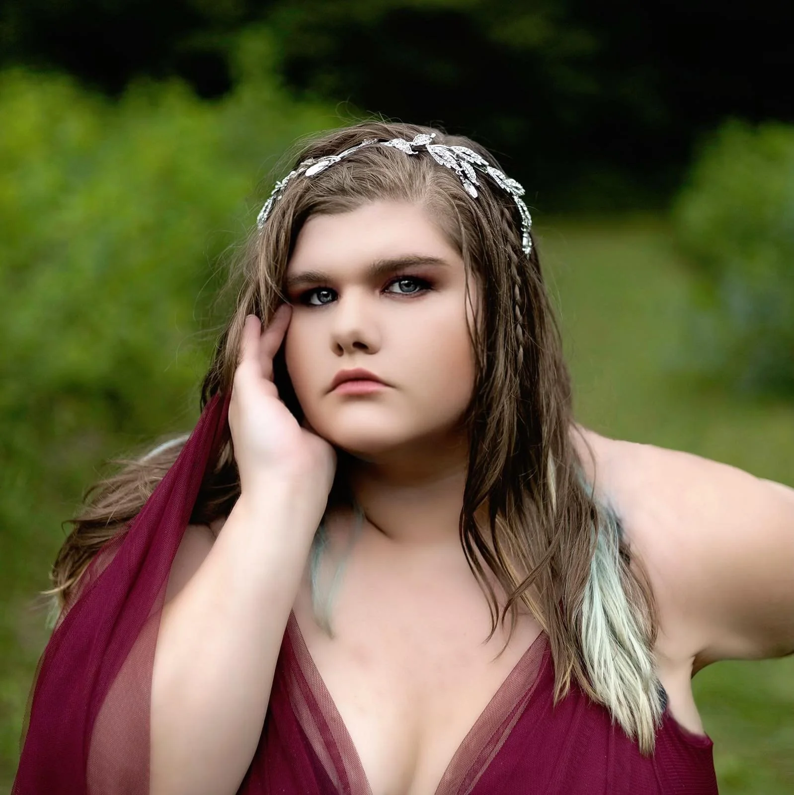 Close-up portrait of a young woman with long brown hair, wearing a silver headband, red dress, and posing outdoors with a blurred green background.