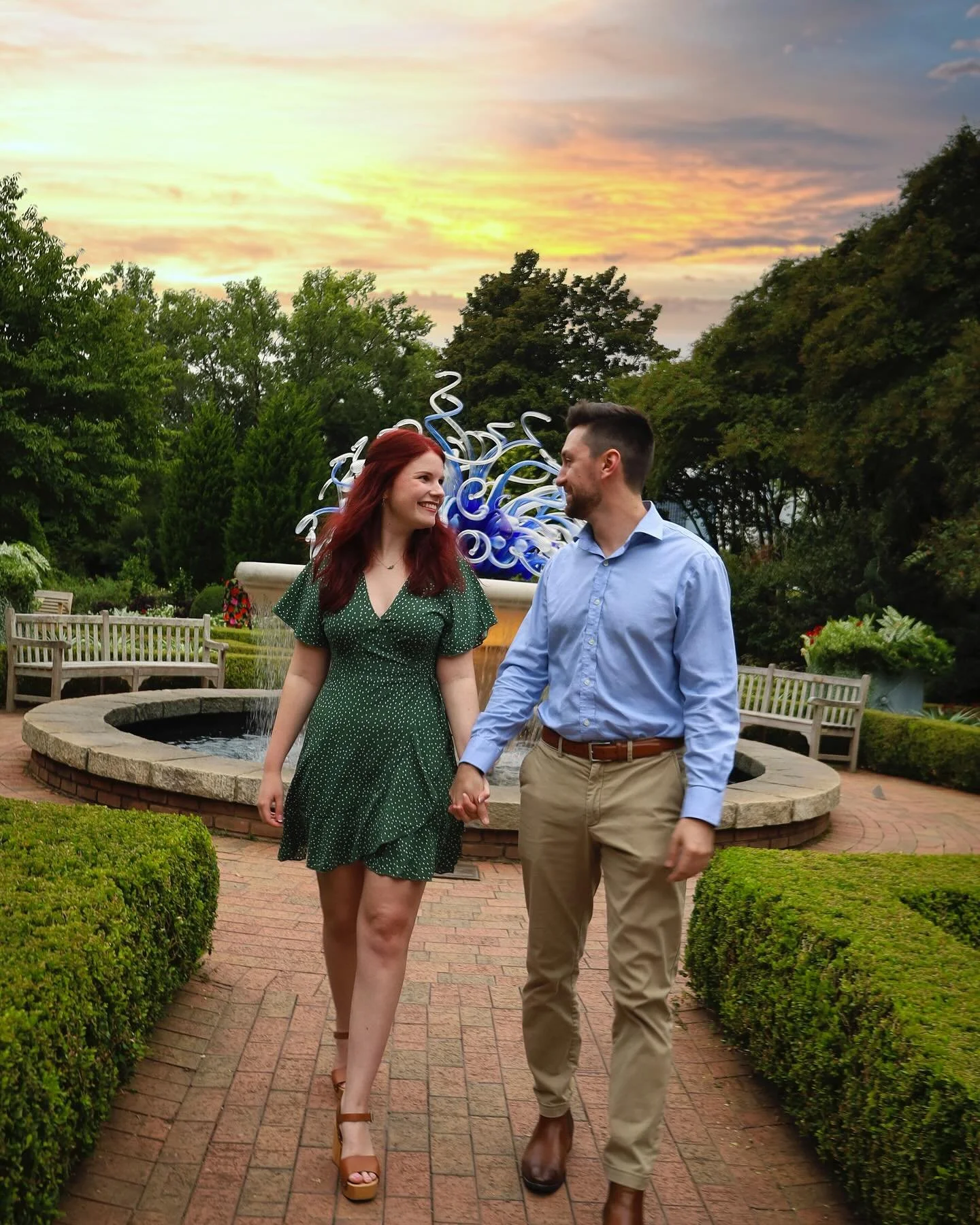 A couple holding hands and smiling at each other while walking through a park with a fountain and colorful glass sculpture in the background, during a sunset.