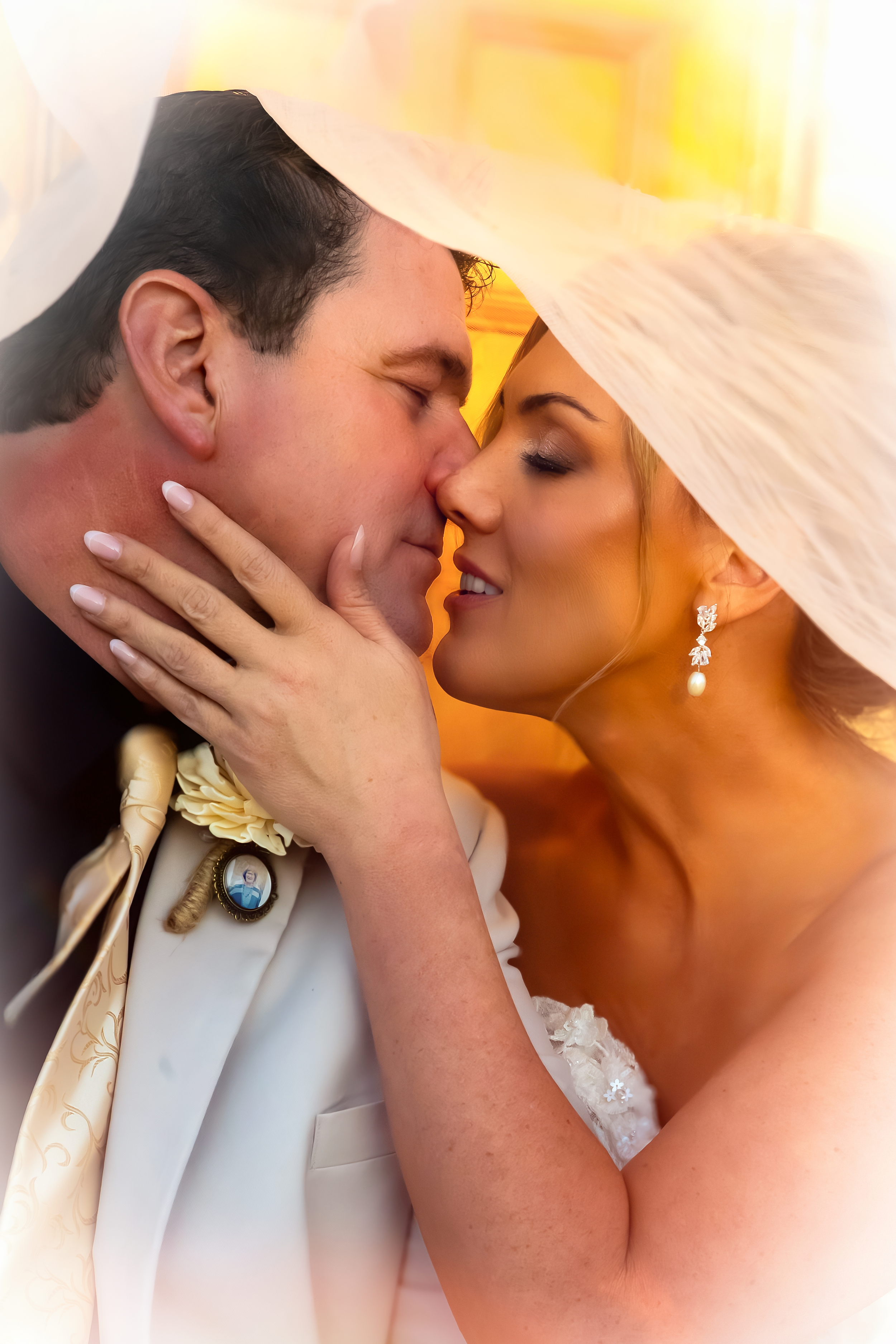 A bride and groom share a close, intimate moment with their foreheads touching, under a large, white veil. The bride wears pearl and diamond earrings and a strapless white wedding gown. The groom wears a light-colored tuxedo with a boutonniere.