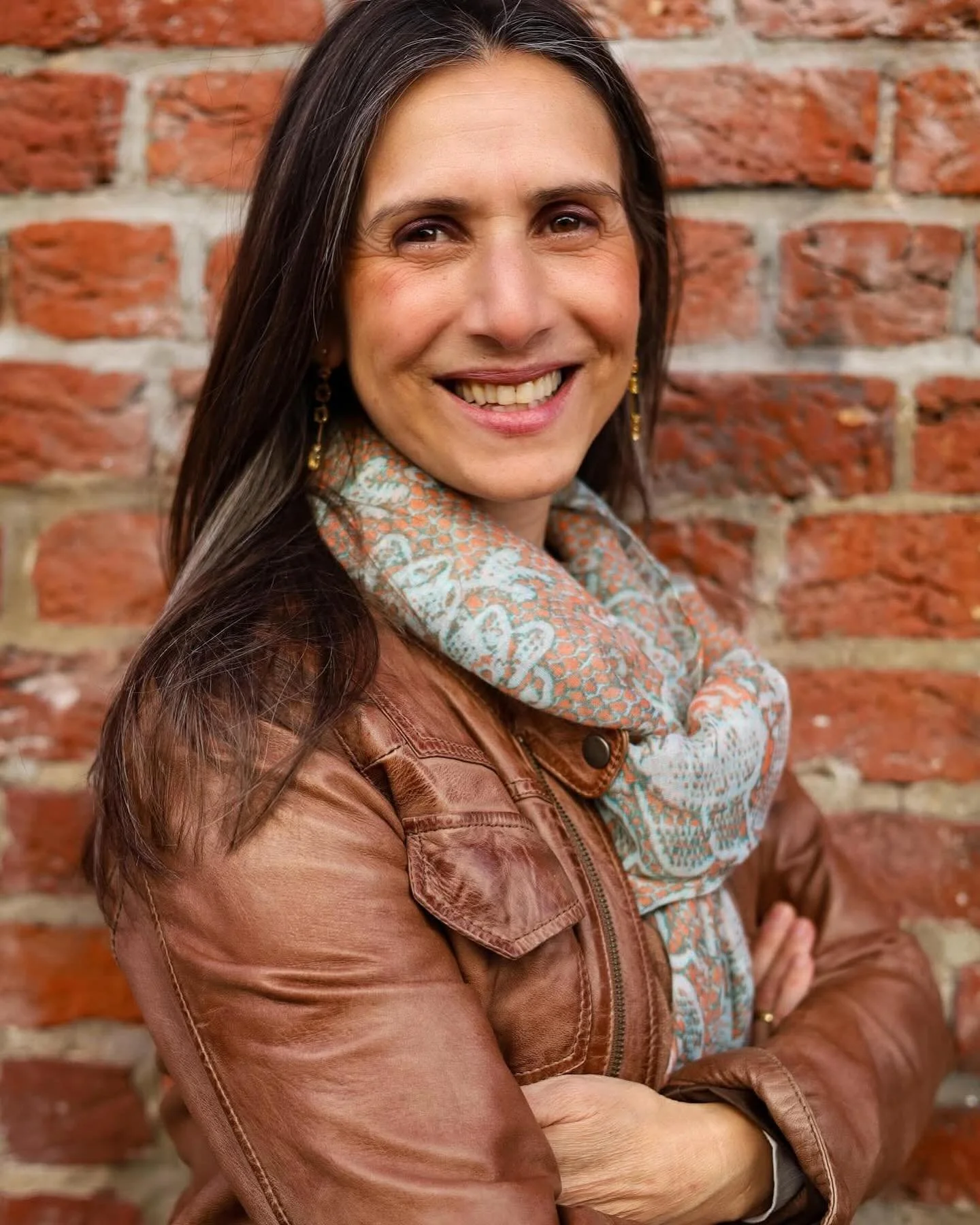 Smiling woman with long dark hair, wearing a patterned scarf and a brown leather jacket, standing against a red brick wall.