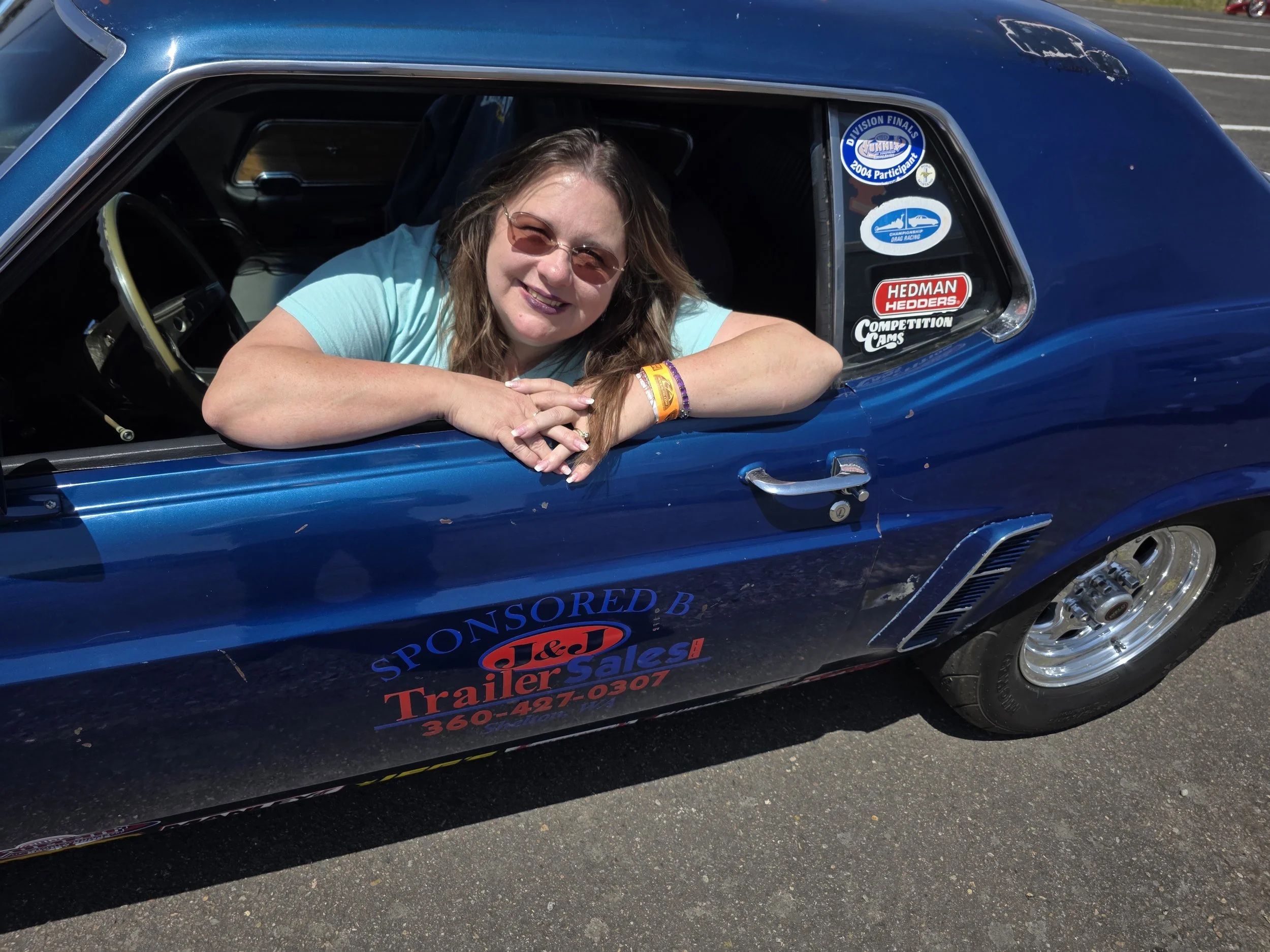 Jahanel leaning out of her 1969 Mustang race car with various sponsor stickers, smiling at the camera.