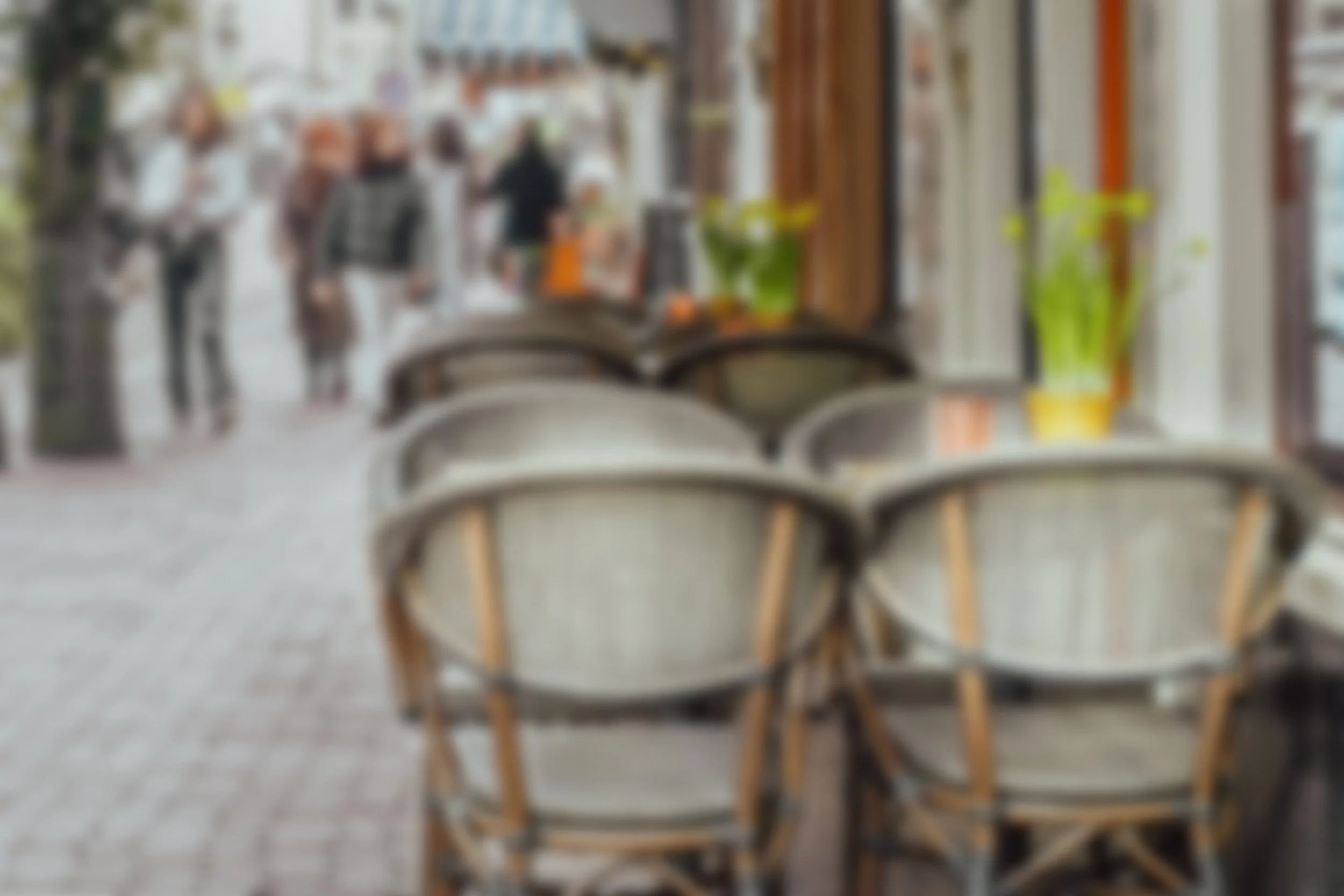 Blurry indoor cafe with empty chairs at a table, windows, and some plants, with people walking outside