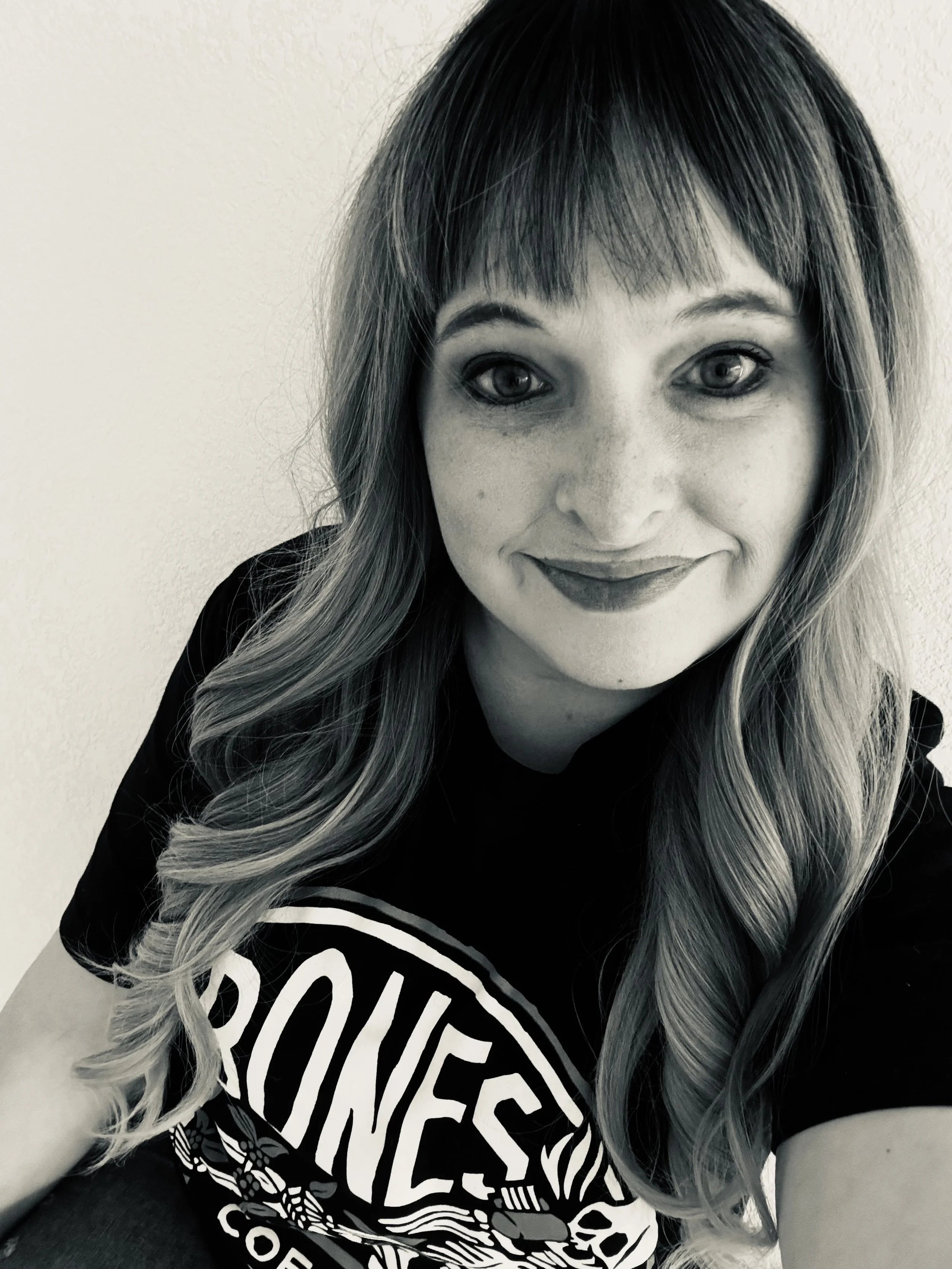 A woman with long wavy hair, wearing a Black Sabbath T-shirt, smiling at the camera, with a plain wall background.