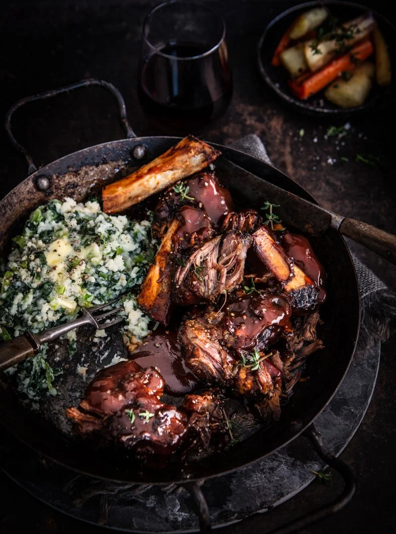 A skillet containing chopped beef short ribs with barbecue sauce, served with mashed potatoes and greens. A glass of red wine and a bowl of roasted vegetables are in the background.