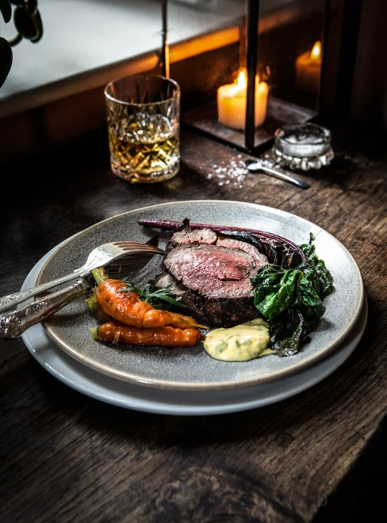 A plate with sliced beef steak, roasted carrots, sautéed greens, and a dollop of creamy sauce, set on a rustic wooden table with a glass of whiskey, lit candles, and a lantern in the background.