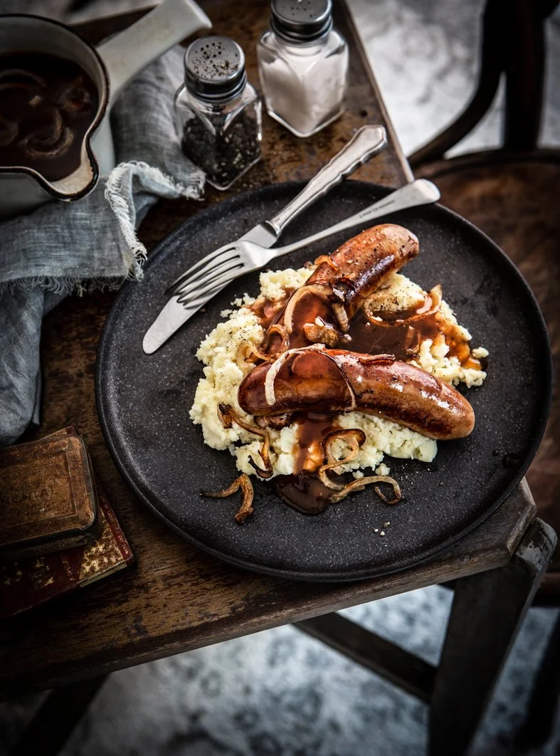 Plate of mashed potatoes topped with sausages, gravy, and fried onions on a black plate.