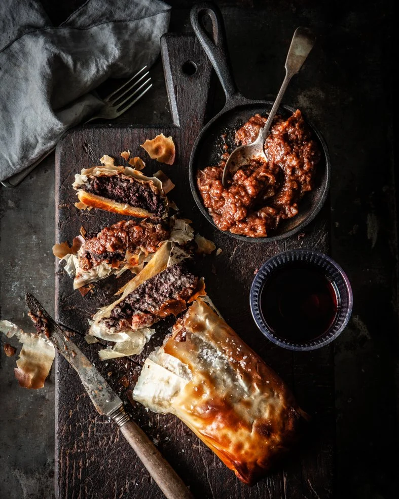 Slices of meat pie with flaky crust on a wooden cutting board, a bowl of beef stew, a glass of red wine, and a fork and knife on a dark surface.