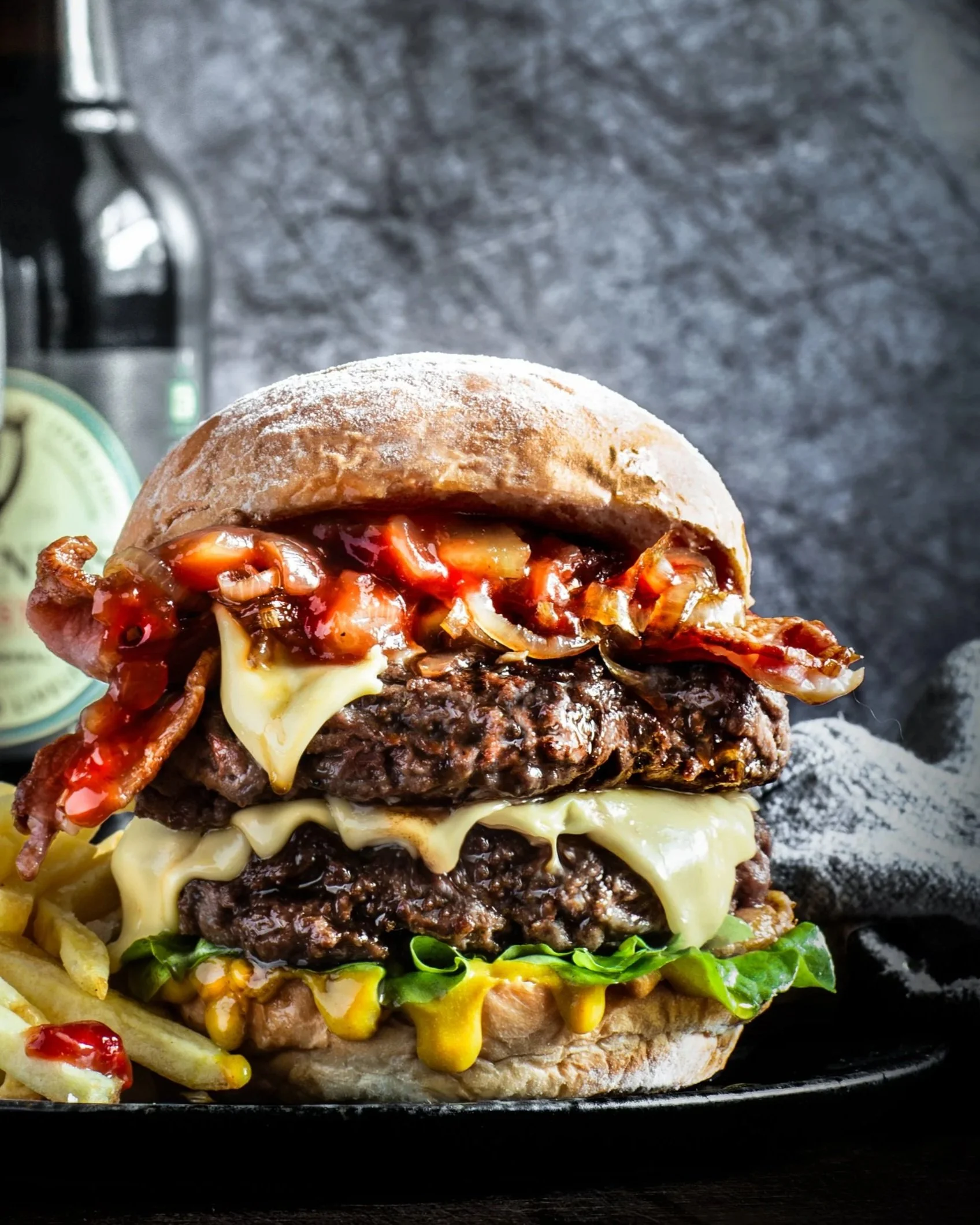 A large guinness cheeseburger with fries and condiments, a glass of dark beer, and a bottle of guinness beer in the background on a dark surface.