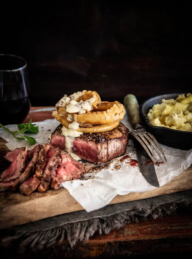 A plate of medium-rare steak topped with fried onion rings and gravy, served with mashed potatoes and a glass of red wine.