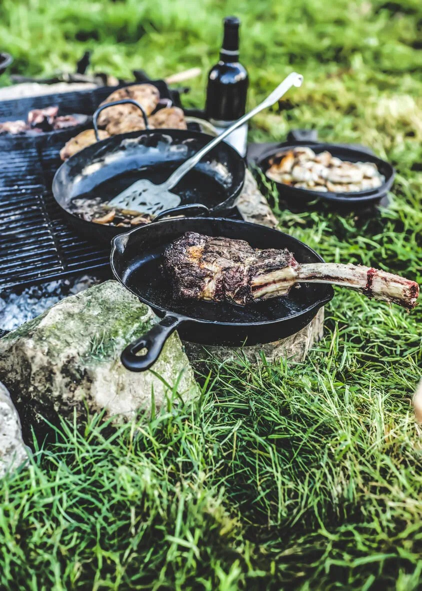 Cast iron skillet with cooked beef short rib, surrounded by other cookware and food on grass outdoors.