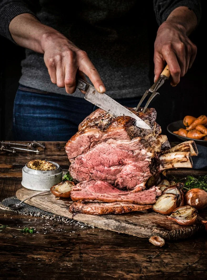 Person carving a large, cooked beef roast on a wooden cutting board, with onions and garlic nearby.