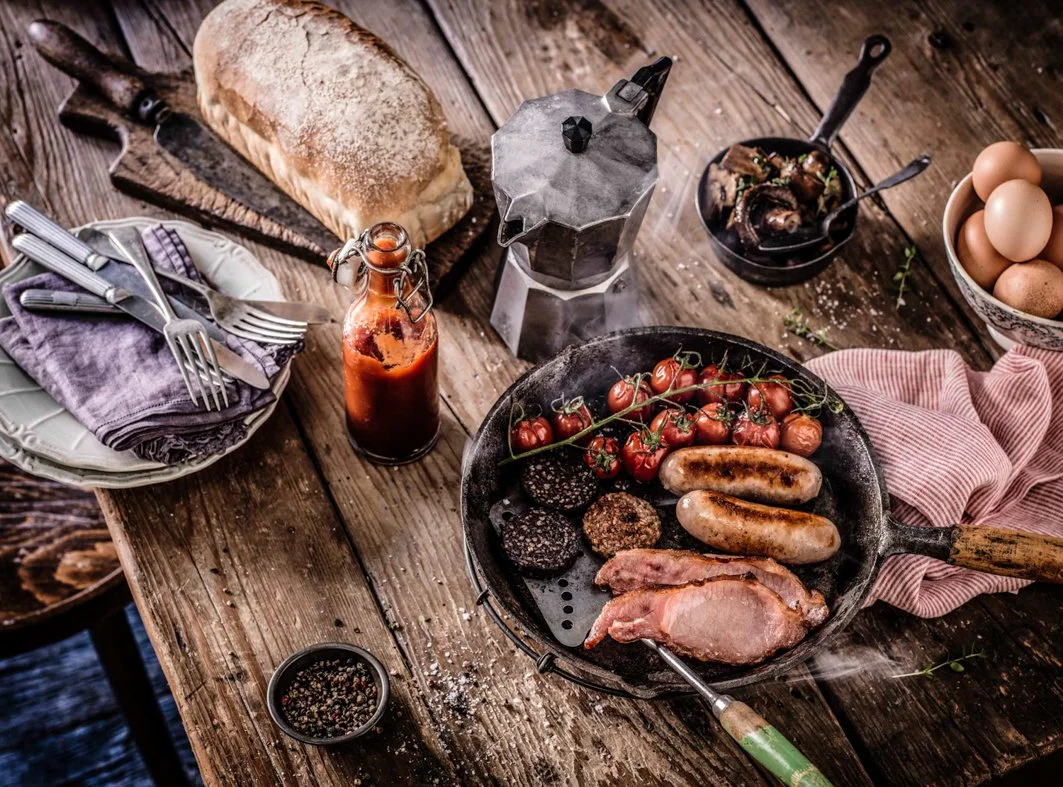 Overview of a rustic wooden table with various breakfast foods and cooking items, including a loaf of bread, a bottle of sauce, a French press, a pan with grilled sausages, cherry tomatoes, ham, and blackened burger patties, a bowl of eggs, and a set