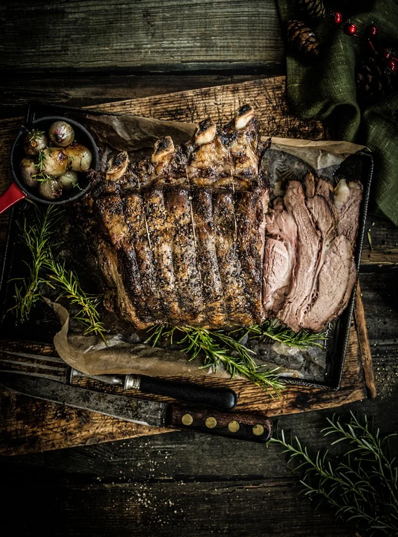Roast beef with sliced beef on the right, roasted onions garnished with herbs in a small pan, and fresh herbs on a rustic wooden table.