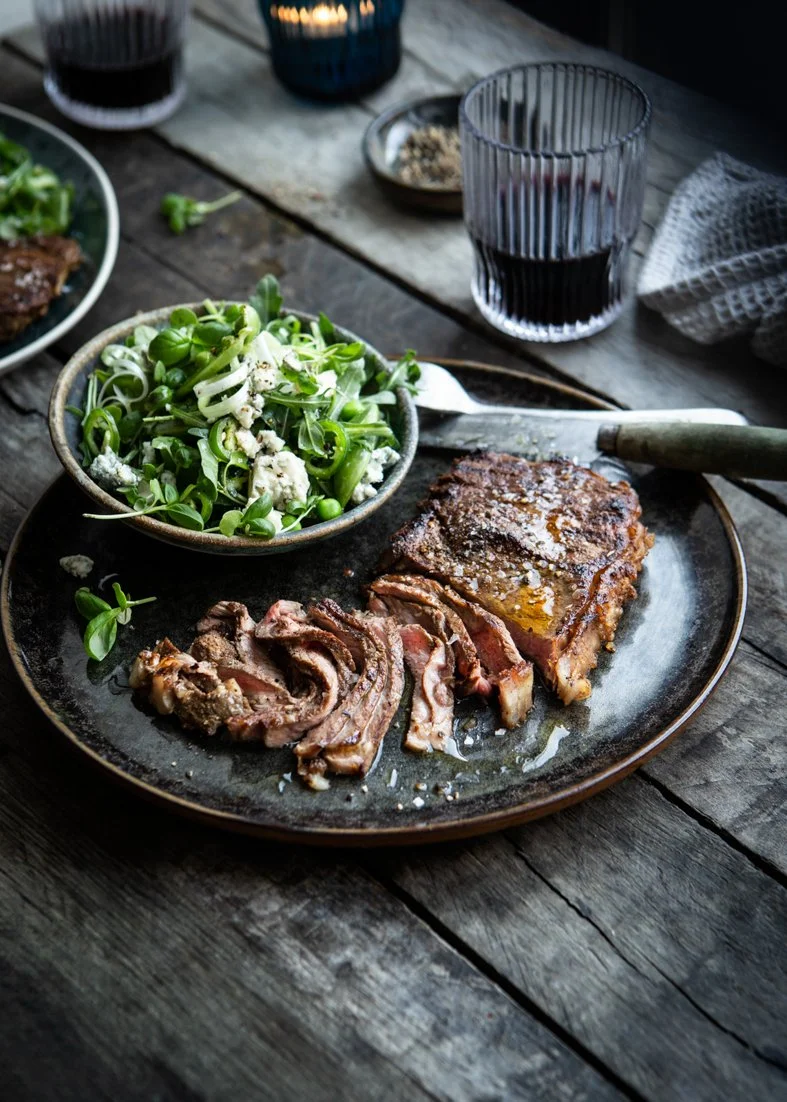 A plate with cooked sliced steak and a salad on a rustic wooden table, with a glass of red wine in the background.