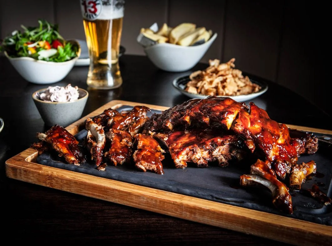 Barbecue ribs with sauce on a wooden platter, sides of salad, coleslaw, potato chips, and a glass of beer on a dark table.