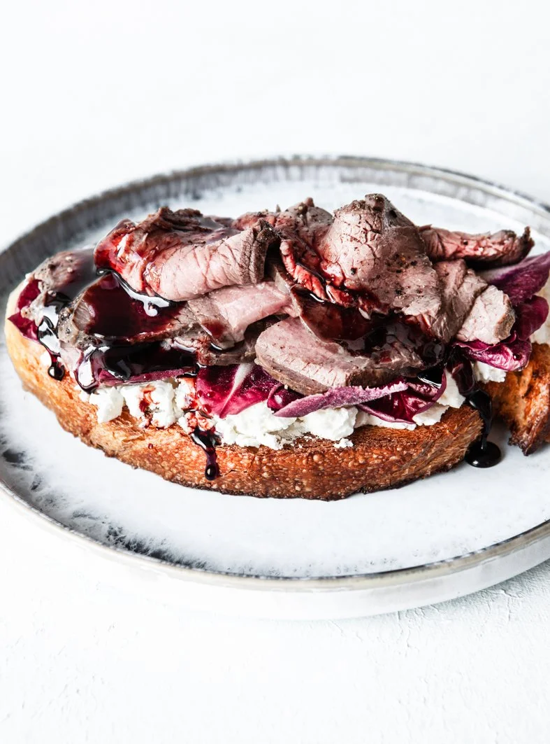 Open-faced sandwich on a toasted bread with sliced roast beef, cheese, radicchio, and a dark sauce drizzle.