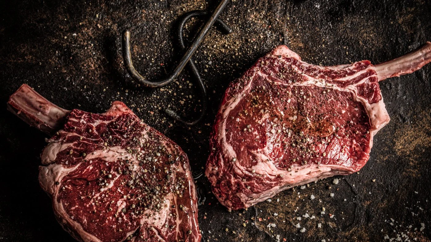 Two raw, seasoned tomahawk steaks on a dark cooking surface with salt, pepper, and a meat fork.