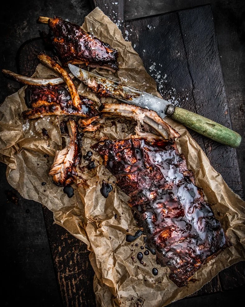 Barbecue ribs with sauce on crumpled brown paper, knife with green handle on dark surface.
