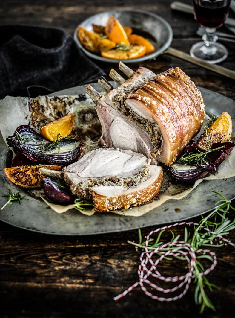 A large roasted pork loin with crispy skin, sliced to show the inside, served on a dark plate with roasted vegetables and garnished with herbs, on a rustic wooden table.