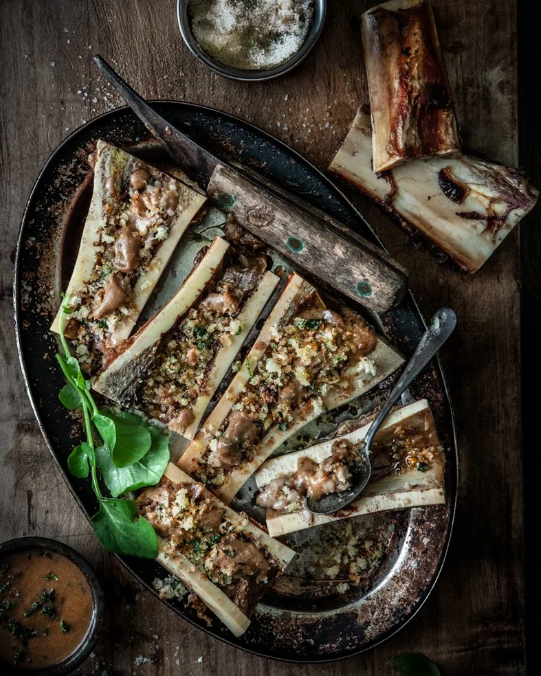 Sliced beef rib on a large metal platter topped with breadcrumbs, garlic, and herbs, served with a side of sauce and garnished with greens, on a rustic wooden table.