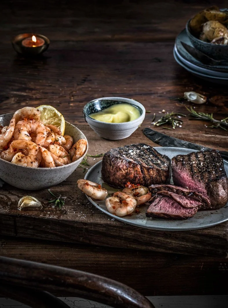 A plate of grilled steak with a side of cooked shrimp, a bowl of dipping sauce, and lemon wedges on a rustic wooden table, with a lit candle in the background.