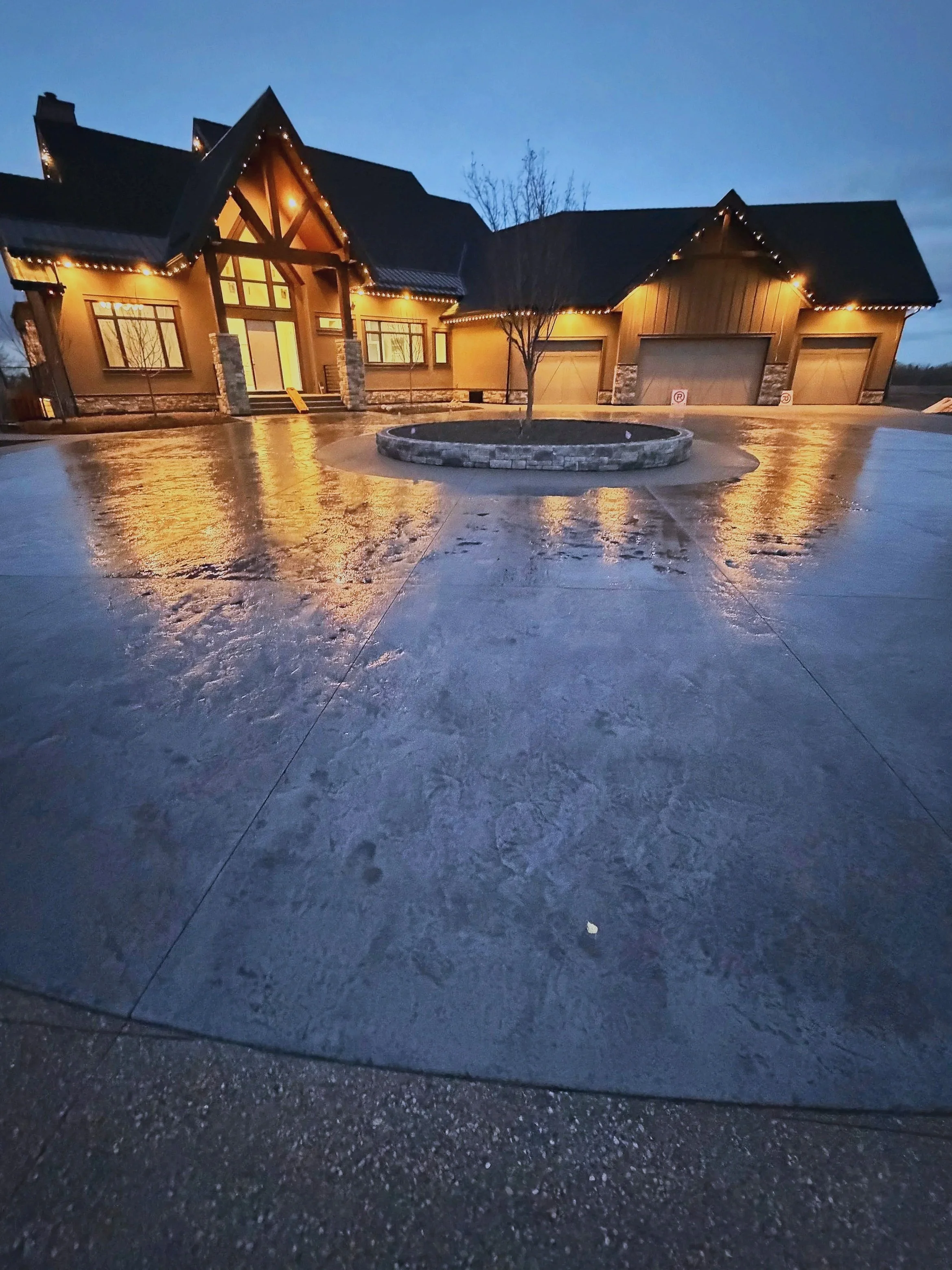 A large house with a steep roof decorated with Christmas lights, illuminated windows, a driveway with wet ice or snow, and a circular raised flower bed with a small tree in the center.