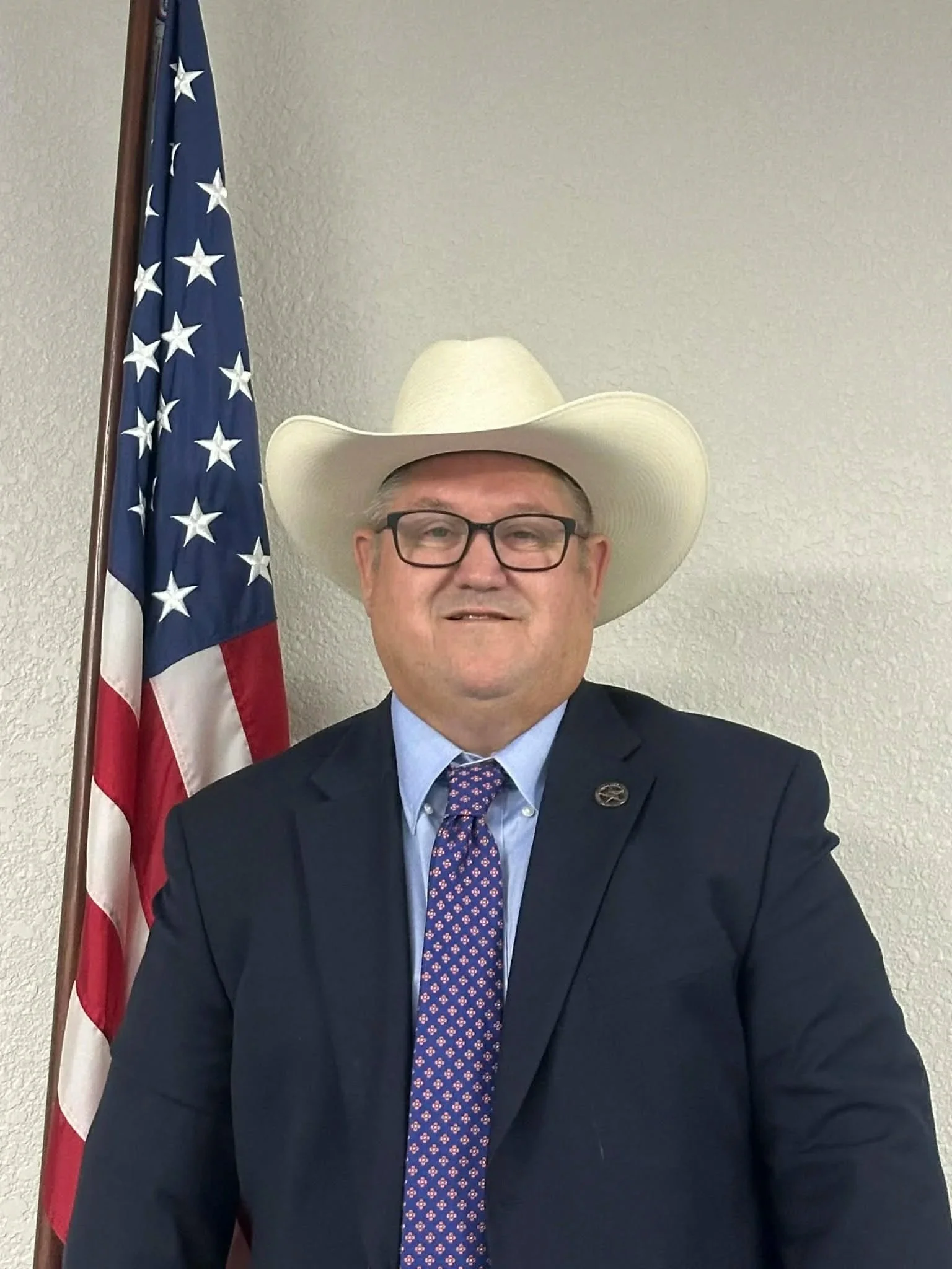 A man in a suit wearing a large cowboy hat and glasses standing in front of an American flag.