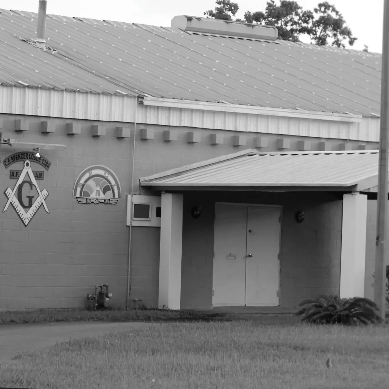 Black and white photo of the exterior of a building with Masonic symbols on the wall, a covered entrance, and some grass in the foreground.