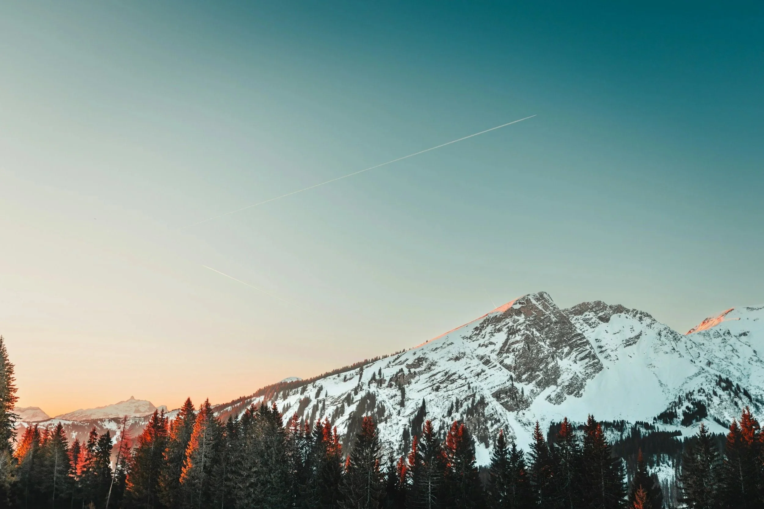 Snow-covered mountain range at sunset with tree line in foreground and contrails in the sky.