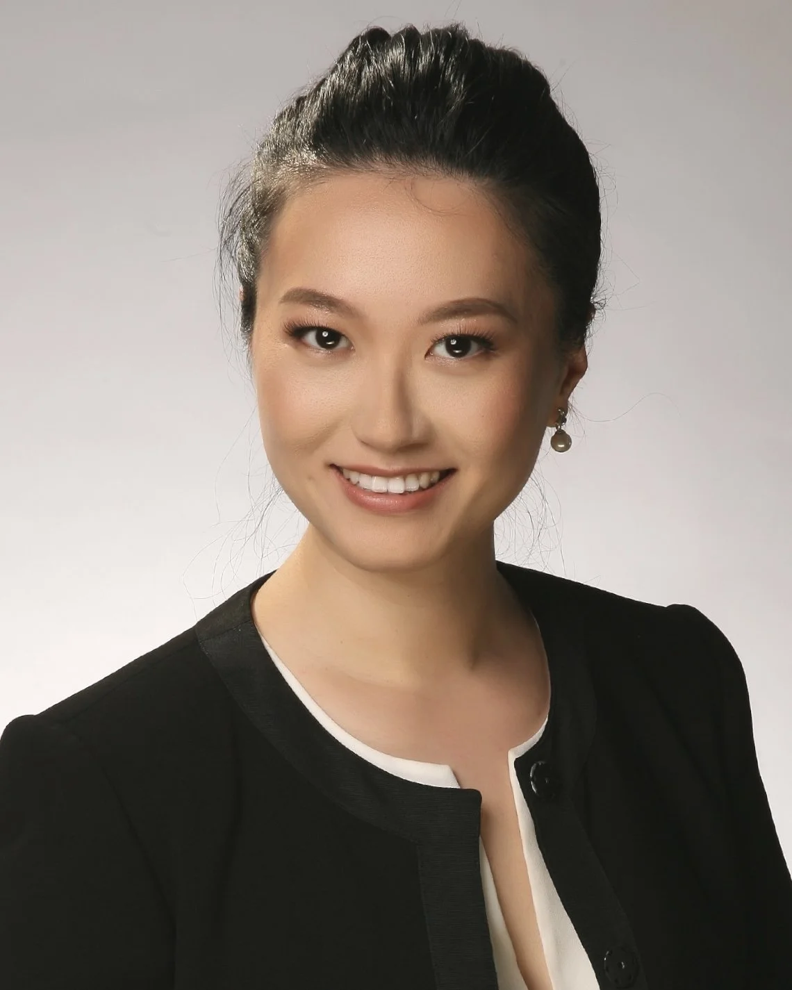 Professional headshot of a smiling Asian woman with dark hair pulled back, wearing a black blazer with white trim and pearl earrings, against a plain background.