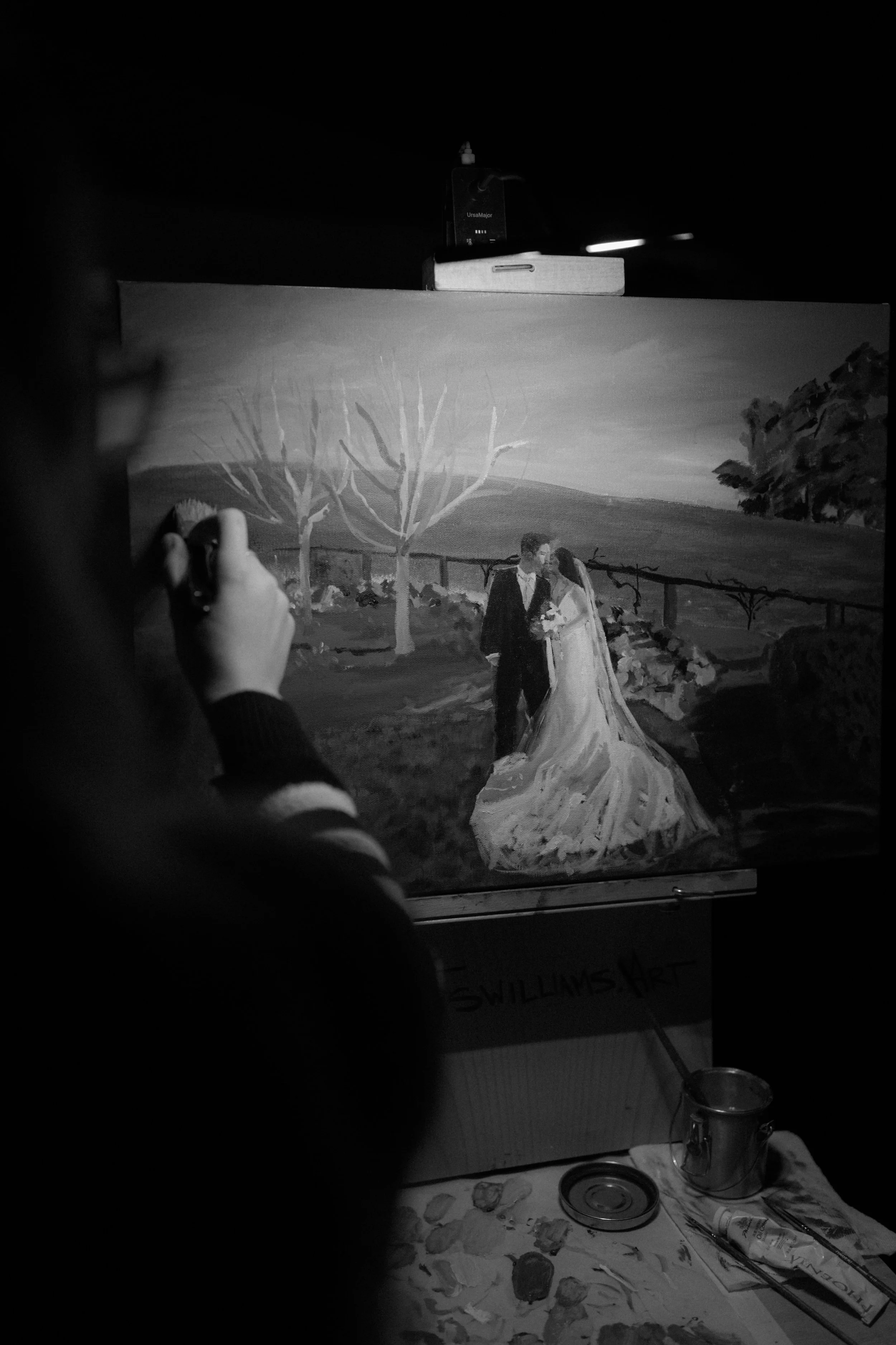 A person painting a monochrome landscape with a couple on their wedding day, showing a bride and groom holding hands and facing each other outdoors with trees and a fence.