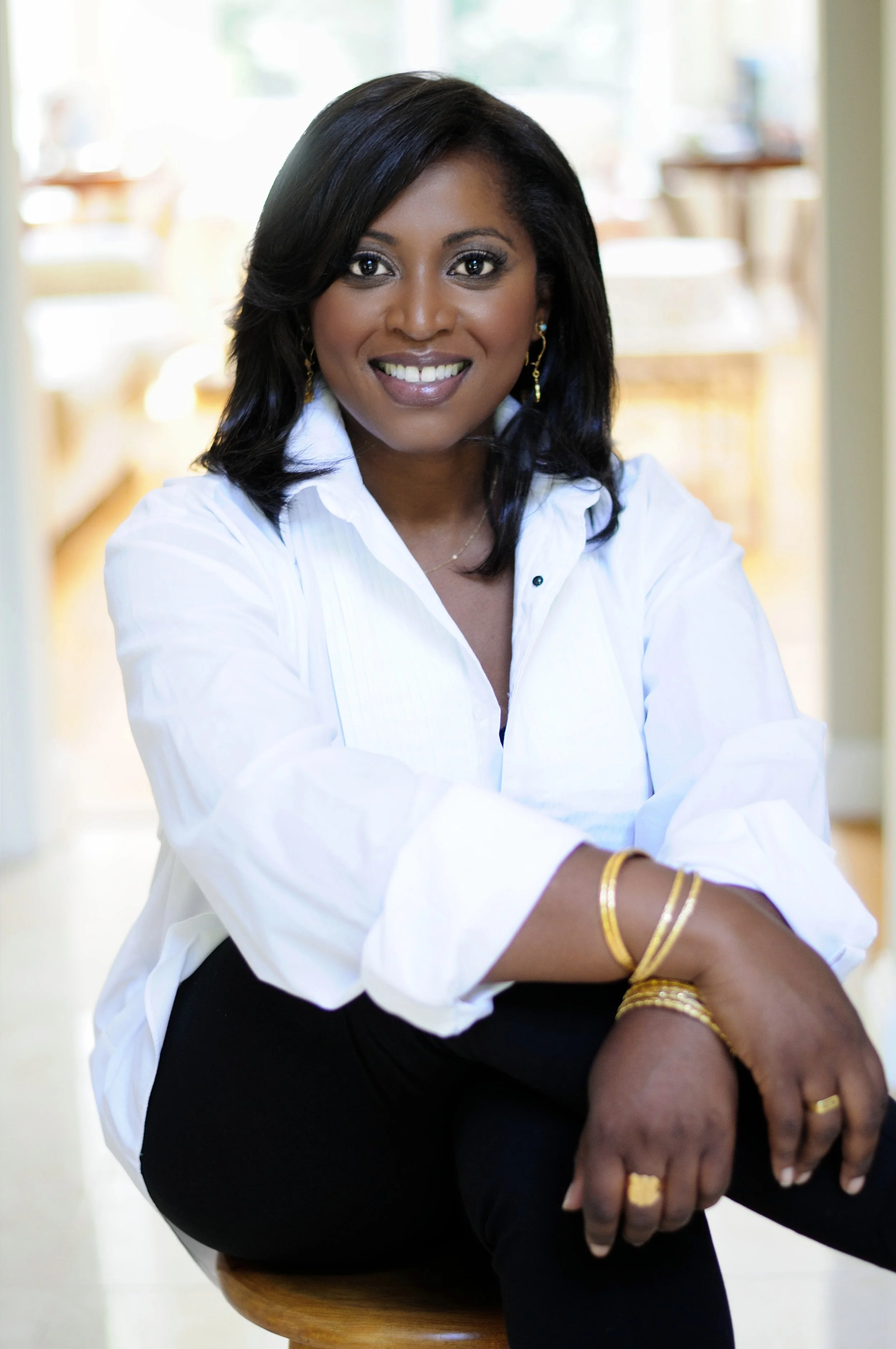 A smiling woman with black hair and brown skin, wearing a white shirt and black pants, sitting on a stool indoors.