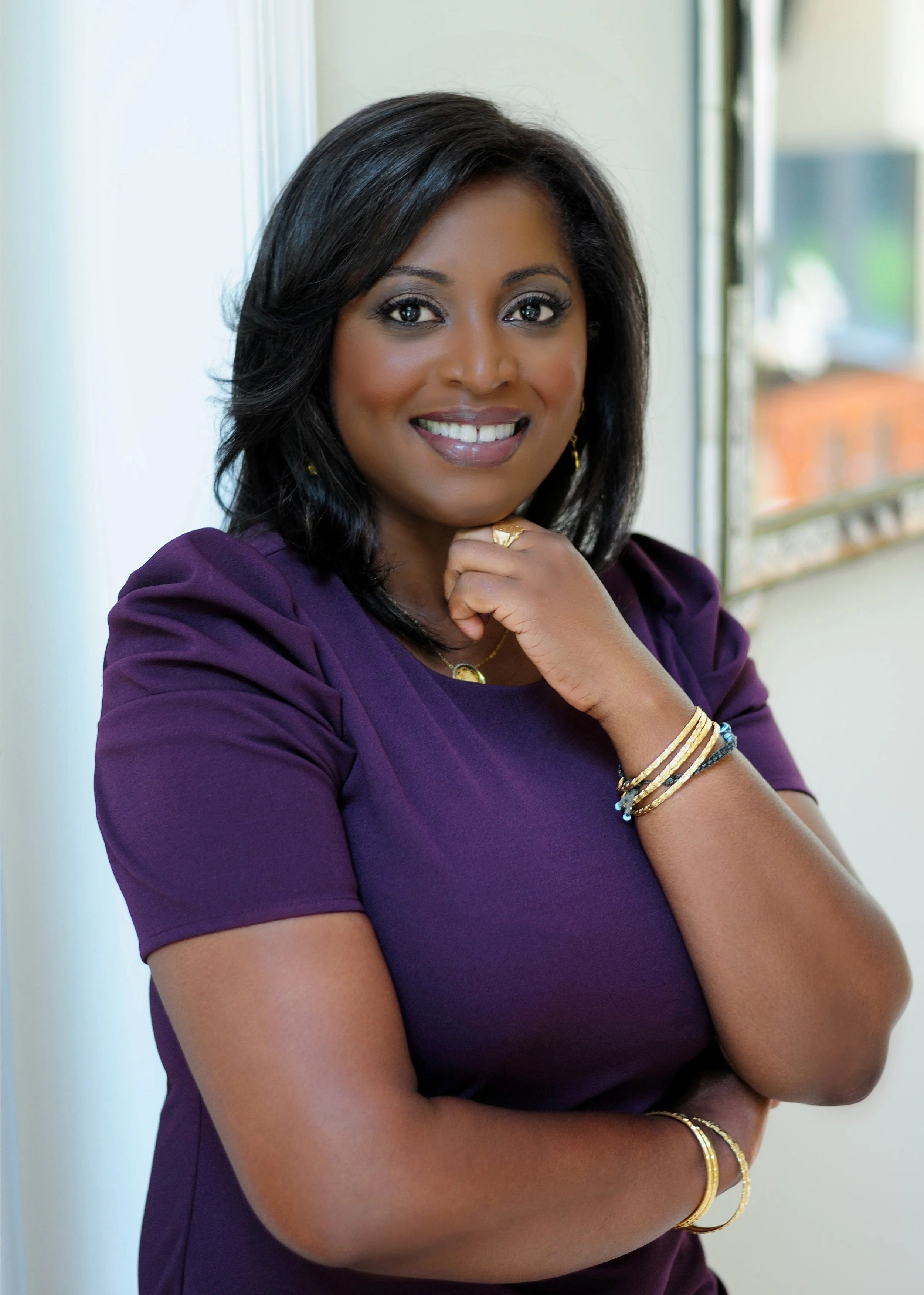 A woman with medium dark skin tone, shoulder-length black hair, wearing a purple short-sleeve dress, smiling, standing next to a window with a view of a balcony and some buildings in the background.