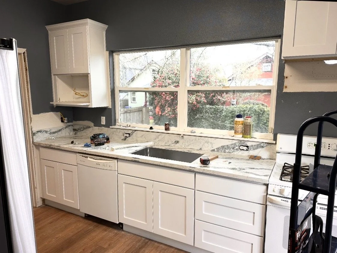 Kitchen under renovation with white cabinets, marble countertop, and large window, showing construction tools and materials.