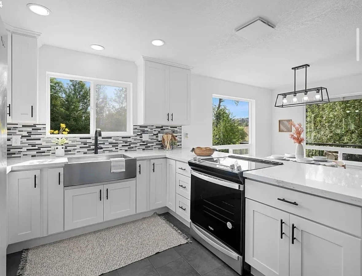 Modern white kitchen with black hardware, subway tile backsplash, stainless steel farm sink, black stove, window views of green trees, and a dining area with a ceiling light fixture.