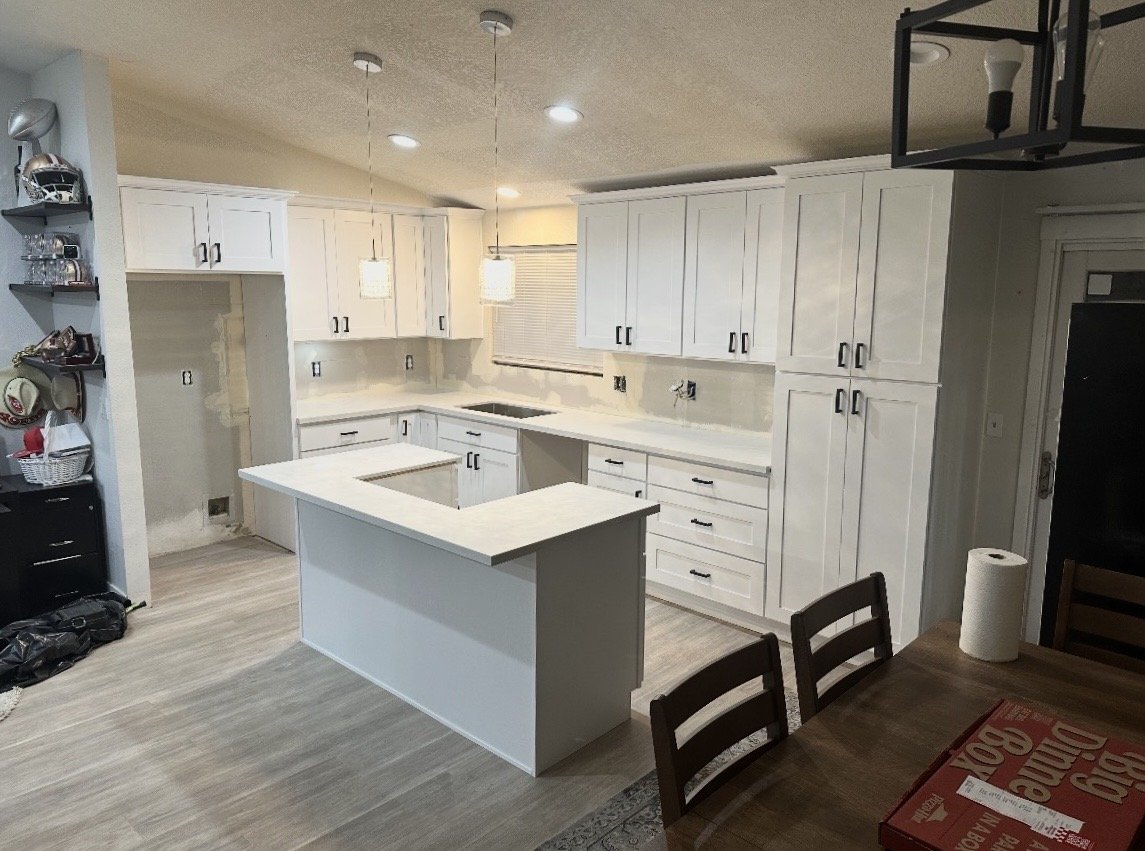 Empty white kitchen with island, upper and lower cabinets, and pendant lighting, adjacent to a dining table with a roll of paper towels and a pizza box.