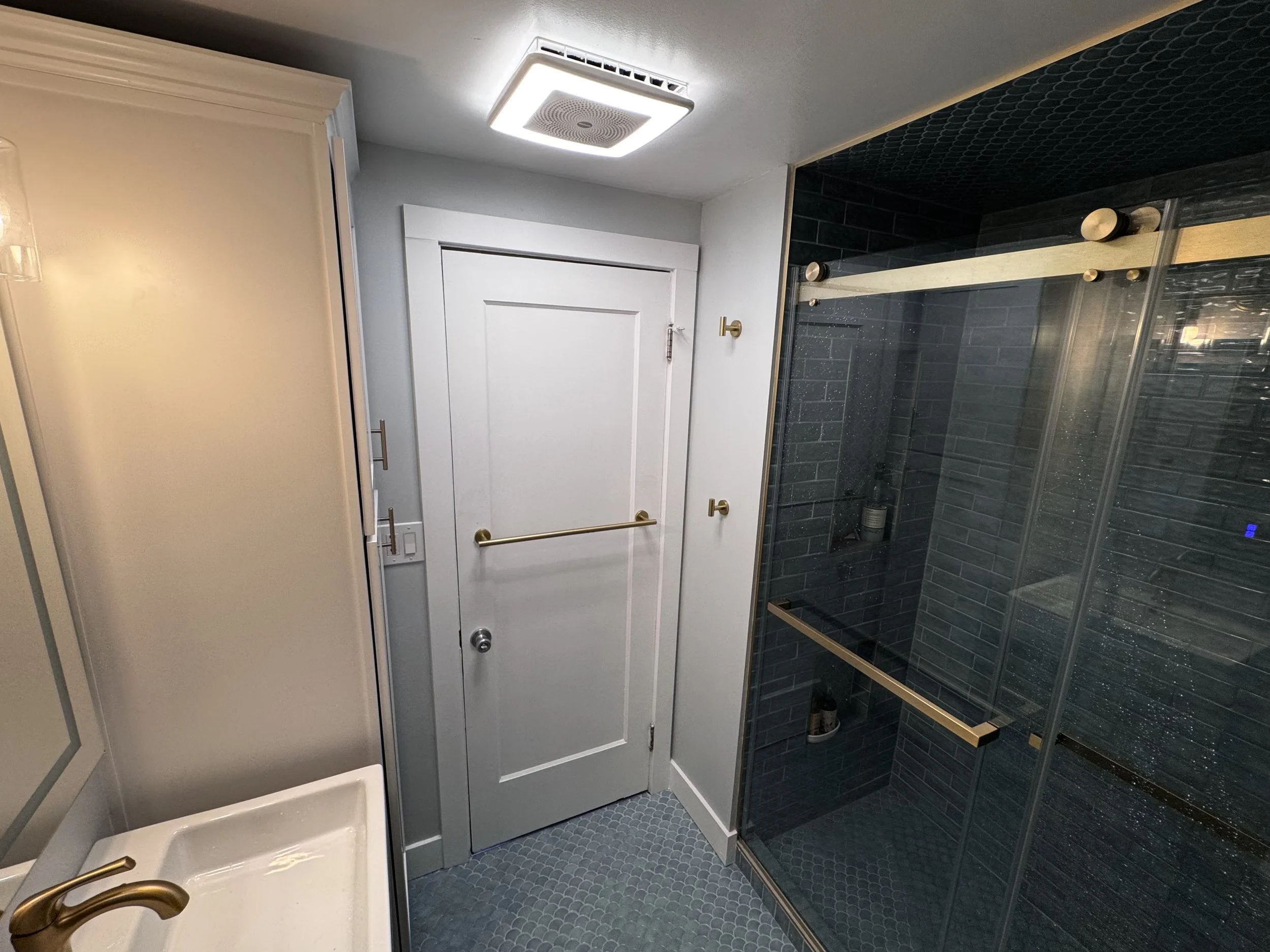 Bathroom with a closed white door, a glass shower enclosure with black brick tile and brass hardware, a white sink with a brass faucet, and blue patterned floor tiles.