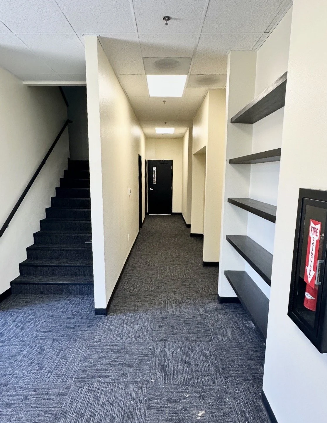 Hallway with black stairs on the left, beige walls, built-in shelves on the right, and a black fire extinguisher case on the wall.