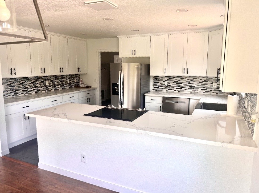 Modern kitchen with white cabinets, a marble countertop island, stainless steel refrigerator, and a black and gray mosaic tile backsplash.
