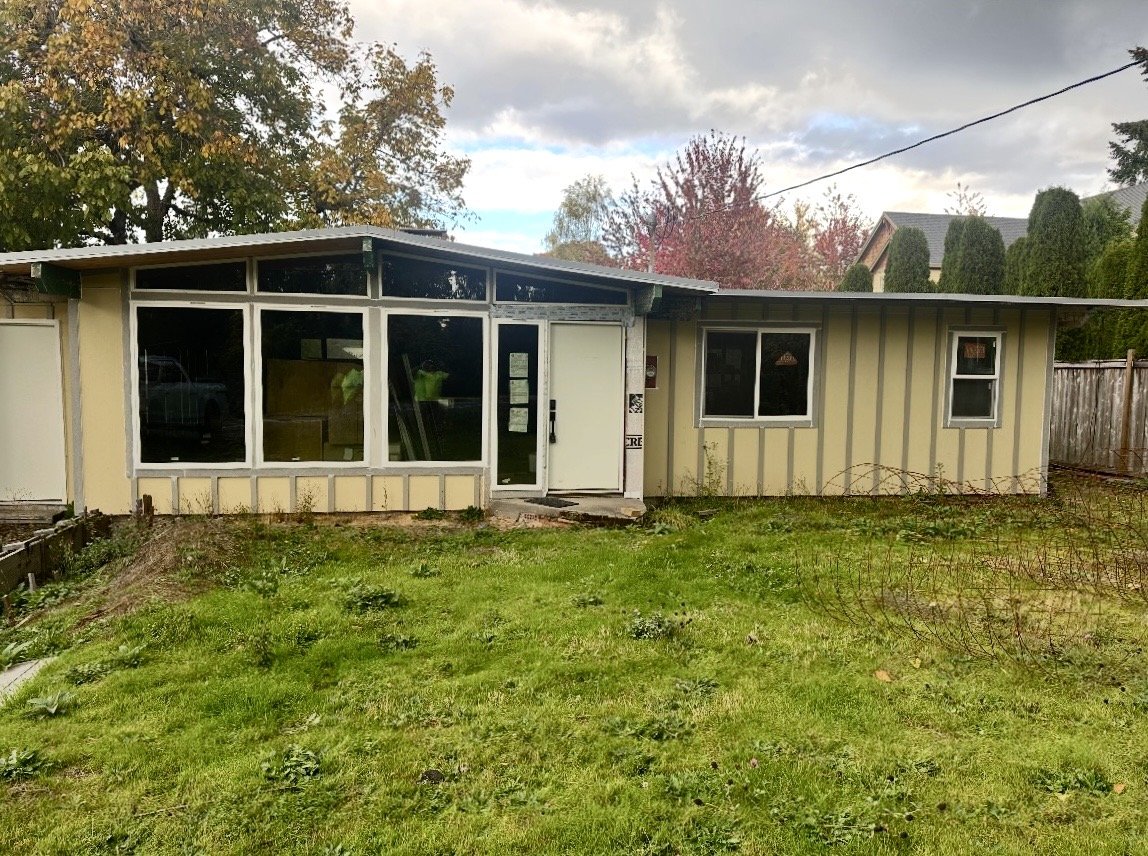 Backyard view of a house with a large glass sunroom, yellow exterior walls with vertical siding, and three windows. The yard has green grass and some small plants, with trees and shrubs in the background, under a partly cloudy sky.