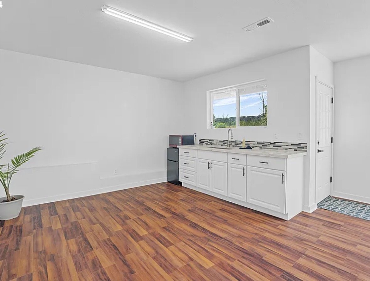 Empty kitchen with white cabinets, a window, a small refrigerator, and wooden flooring.