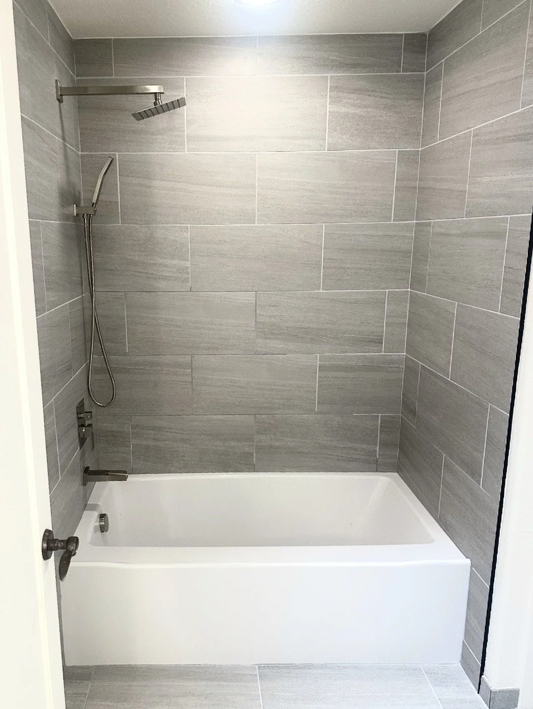 A modern walk-in shower with gray tile walls, a rain showerhead, a handheld shower, and a white bathtub.
