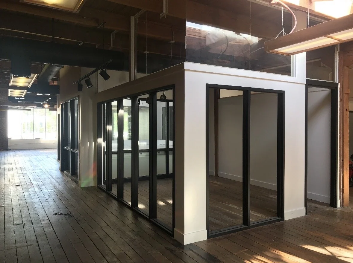 An office interior with small glass-walled rooms or partitions, wooden flooring, and modern lighting fixtures.