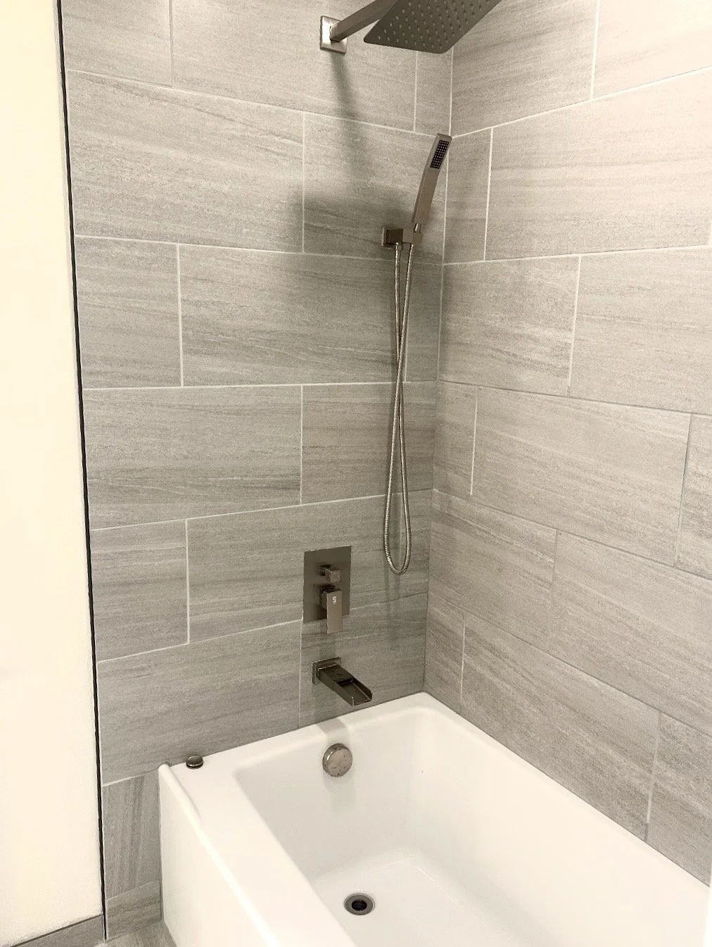 Modern bathroom shower with gray tiled walls, a large square showerhead, a handheld showerhead, and a bathtub with a silver drain.
